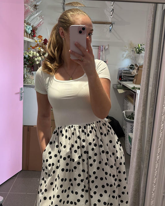 A woman takes a mirror selfie in a room, wearing a white short-sleeve scoop-neck top and a white skirt with black polka dots.