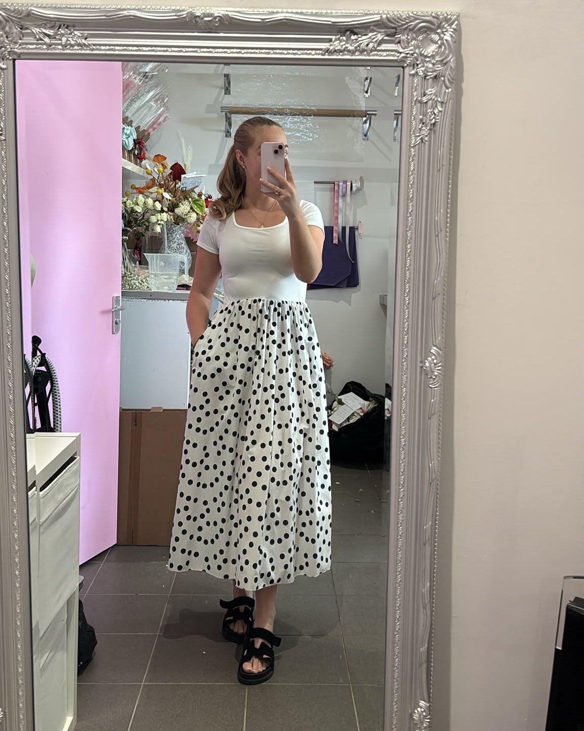 A woman takes a mirror selfie in a boutique showing a white short-sleeve top and a white midi skirt with black polka dots. The boutique has a large ornate silver mirror, a pink wall, and a shelf with flowers and products.