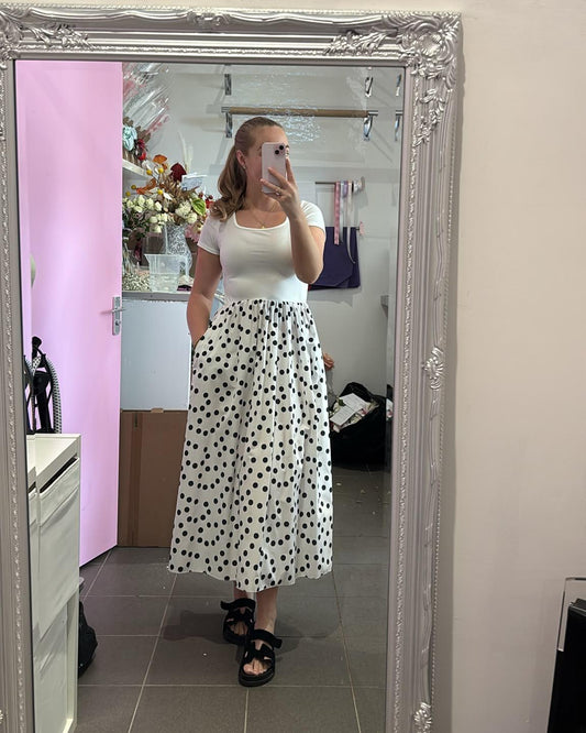 A woman takes a mirror selfie in a boutique showing a white short-sleeve top and a white midi skirt with black polka dots. The boutique has a large ornate silver mirror, a pink wall, and a shelf with flowers and products.