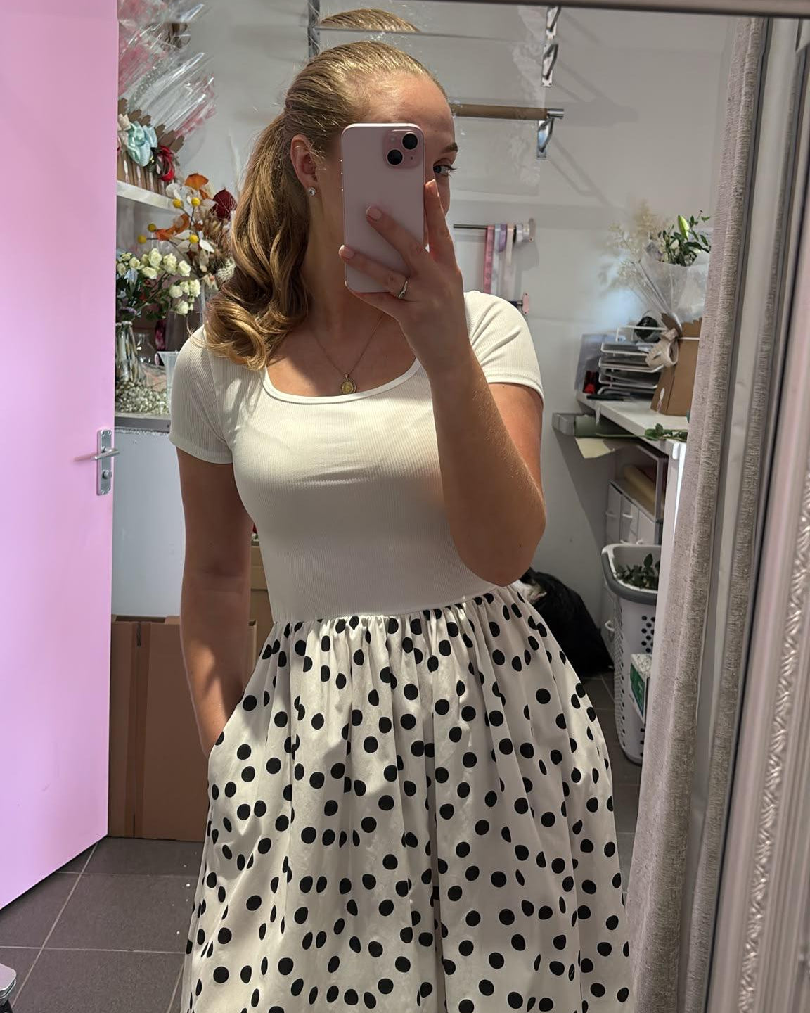 A woman takes a mirror selfie in a room, wearing a white short-sleeve scoop-neck top and a white skirt with black polka dots.